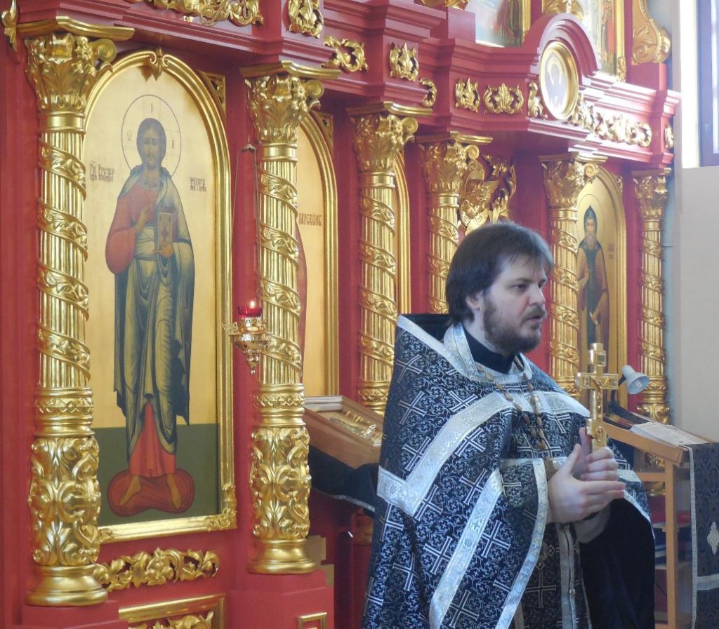 Настоятель храма протоиерей Вадим Антипин стоит на фоне иконостаса во время проповеди с напрестольным храмом в руках.