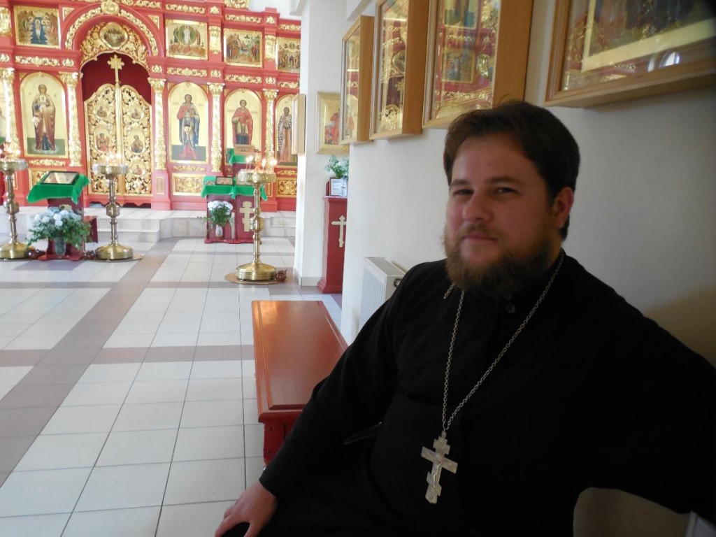 отец Олег Смирнов сидит на лавке в храме Пантелеимона, смотрит в камеру и улыбается.