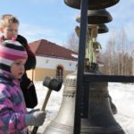 Ребенок дошкольного возраста стоит рядом с передвижной звоннице на прихрамовой территории и ударяет киянкой в большой колокол